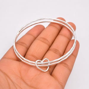 May include: A silver bangle bracelet featuring a double-layered design with a small, open heart-shaped charm linked to the two thin, polished metallic bands. The circular jewelry piece has a smooth, reflective surface and a simple, clean construction.