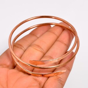 May include: A copper-colored metal bangle bracelet featuring a multi-coil design. The bracelet is constructed from a single continuous wire that wraps around to form three distinct circular layers, with smooth, rounded ends at each terminal point.