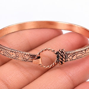 May include: A copper bangle bracelet featuring an embossed floral pattern along the band. The bracelet closes with a twisted metal ring mechanism that connects the two ends, creating a circular shape with a metallic reddish-brown finish.