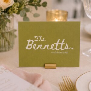 May include: A rectangular olive green card held by a small gold metal stand. The card features white text that reads The Bennetts. with a small heart icon and the text established 2026 printed in a smaller font underneath the main name.