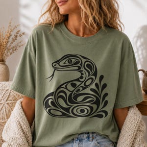 Może przedstawiać: Oliwkowozielony t-shirt z krótkim rękawem z dużym czarnym nadrukiem graficznym przedstawiającym zwiniętego węża o stylizowanych, płynnych liniach i rozdwojonym języku. Koszulka ma okrągły dekolt i luźny krój wykonany z miękkiej, spranego materiału.