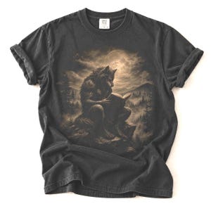 May include: A charcoal grey short-sleeve t-shirt featuring a sepia-toned graphic of a werewolf sitting on a rocky cliff, reading an open book under a full moon. The background depicts a dense forest landscape with mountains under a cloudy sky.