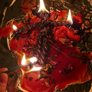 May include: A dark twisted candle shaped like a human figure is burning on a sheet of aluminum foil. The candle is surrounded by melted red wax, scattered dried flower petals, and herbs. Multiple flames are visible on the candle and wax.