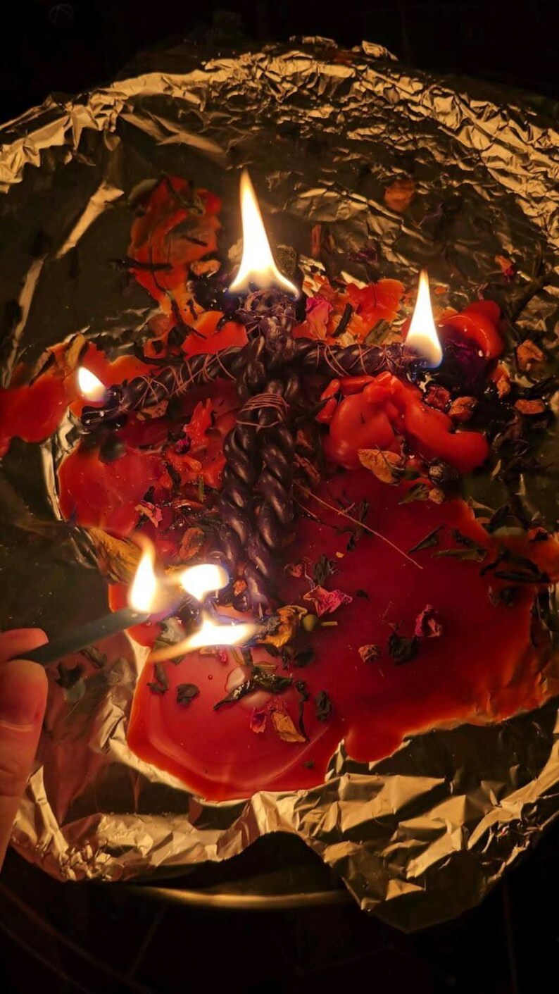 May include: A dark twisted candle shaped like a human figure is burning on a sheet of aluminum foil. The candle is surrounded by melted red wax, scattered dried flower petals, and herbs. Multiple flames are visible on the candle and wax.