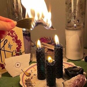 May include: Three black honeycomb-textured pillar candles burning on a white plate with red trim. The plate holds dried herbs, two blue glass stones, a black stone, and an amethyst cluster. A burning green leaf with the text I am all is held above.