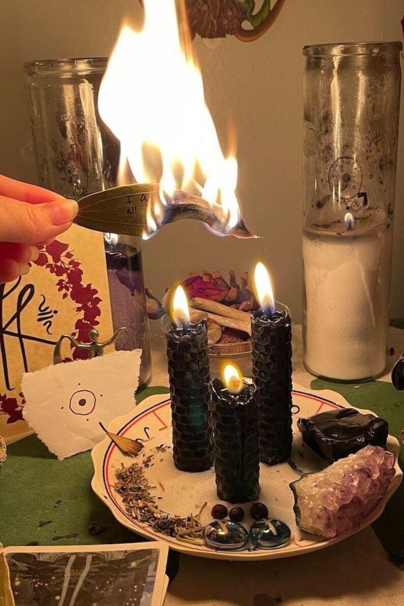 May include: Three black honeycomb-textured pillar candles burning on a white plate with red trim. The plate holds dried herbs, two blue glass stones, a black stone, and an amethyst cluster. A burning green leaf with the text I am all is held above.