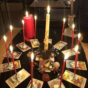 May include: A circular arrangement of tarot cards on a black cloth, each featuring a lit red candle secured with aluminum foil. A central dark cup holds a tall cream candle with gold symbols, surrounded by small crystals and a miniature elephant figurine.