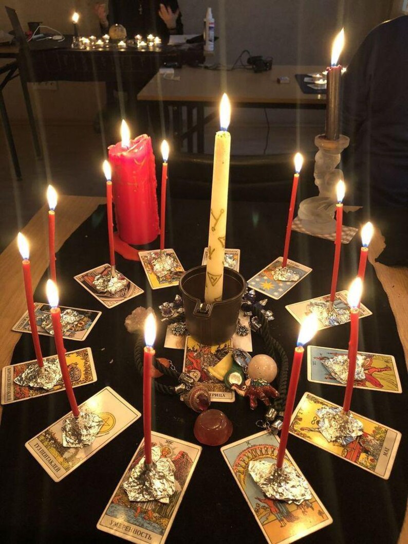 May include: A circular arrangement of tarot cards on a black cloth, each featuring a lit red candle secured with aluminum foil. A central dark cup holds a tall cream candle with gold symbols, surrounded by small crystals and a miniature elephant figurine.