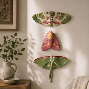 May include: Three wall-mounted moth sculptures arranged vertically. The top moth is cream with green and pink accents. The middle moth is pink with yellow markings. The bottom moth is green with long, trailing tails and small red eye-spot details.