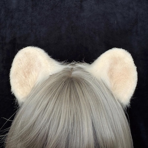 May include: A pair of cream-coloured, plush, rounded animal ears attached to a headband. The ears are covered in soft, short faux fur and are positioned on top of a head featuring straight, light ash-grey hair that is parted in the centre.