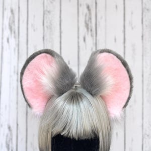Op de afbeelding: Een paar pluche, afgeronde dierenoren bevestigd aan een haarband. De oren zijn voorzien van zacht, grijs imitatiebont aan de buitenranden en een lichtroze, pluizige binnenkant. De haarband is geplaatst op een etalagepophoofd met grijs haar.