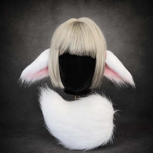 Op de afbeelding: Een korte blonde bob-pruik met een rechte pony, gecombineerd met twee grote, puntige, witte en roze imitatiebont dierenoren aan de zijkanten. Een dikke, pluizige witte imitatiebont staart is onder de pruik op een zwarte standaard geplaatst.