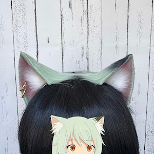 Può includere: Un paio di orecchie da gatto in pelliccia sintetica color verde salvia pallido con interno bianco. Le orecchie presentano due piccoli anelli dorati sul lato destro e sono posizionate sopra una testa con capelli neri lisci, con un'illustrazione.