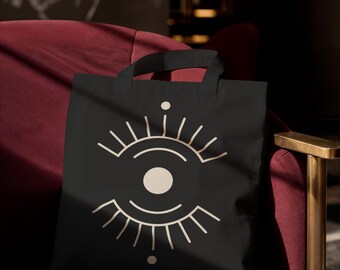 May include: A black tote bag featuring a centered, cream-colored graphic design of an eye with stylized eyelashes, a central pupil, and small circular accents positioned above and below the iris, all rendered in clean, minimalist line art.