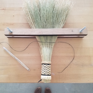 Puede incluir: Una escoba hecha a mano con un mango de madera y una base de paja tejida. Las cerdas de la escoba están hechas de hierba seca y se aseguran con un soporte de madera y sujetadores metálicos. También se ve una herramienta de madera e hilo negro.