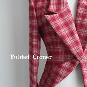 Puede incluir: Una americana de cuadros rojos y grises con mangas largas, solapa de muesca y corte estructurado. La tela muestra un patrón de tartán tejido en tonos carmesí, escarlata y gris claro. El texto "Folded Corner" está impreso en color blanco.