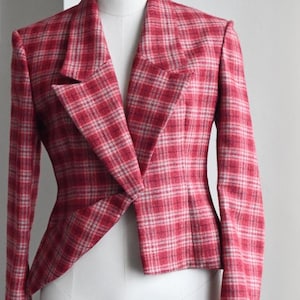 May include: A long-sleeve blazer featuring a red and white plaid pattern. The garment has a notched lapel collar, structured shoulders, and a single-button closure at the waist. The hem is slightly asymmetrical, and the fabric has a textured finish.