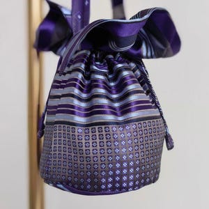Puede incluir: Una bolsa con cordón hecha de corbatas reutilizadas, con una paleta de colores vibrantes en morado y azul. La sección superior muestra rayas horizontales, mientras que la parte inferior presenta un patrón de cuadrícula con motivos cuadrados.