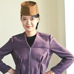 May include: A brown pillbox hat with a flat top and a structured cylindrical shape, paired with a purple long-sleeved jacket featuring a V-neckline, a central silver zip closure, and subtle gathered detailing at the waistline.