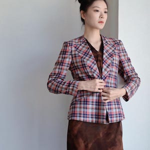 May include: A long-sleeved blazer featuring a plaid pattern in shades of light blue, dark blue, and coral red. The blazer has a collarless neckline and a single-button closure. It is worn over a dark brown dress with a subtle, mottled pattern.