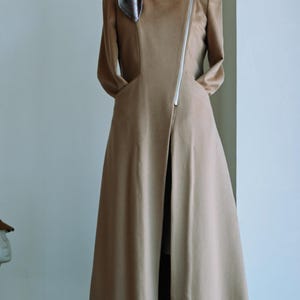 May include: A long, tan-colored coat featuring a square neckline, long sleeves, and an asymmetrical silver-toned zipper closure that runs diagonally across the front. A decorative, iridescent, leaf-shaped brooch is pinned to the upper right chest.