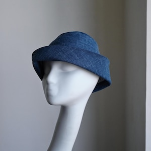 Może przedstawiać: Niebieski kapelusz typu bucket hat o teksturowanym, tkanym wyglądzie z szerokim, opadającym rondem. Kapelusz ma zaokrągloną główkę i jest prezentowany na gładkiej, białej głowie manekina, eksponując krzyżowy wzór materiału oraz głęboki odcień.