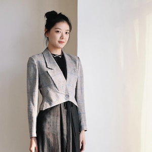 Peut inclure: Un blazer court à motif écossais beige, gris et bleu, doté de manches longues, d'un revers cranté et d'une fermeture à un seul bouton à la taille. Le blazer est porté sur un haut noir et une longue jupe noire fluide à la texture subtile.