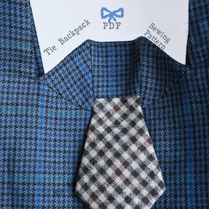 May include: A blue and black houndstooth fabric background with a white paper label featuring a blue bow icon and the text Tie Backpack PDF Sewing Pattern. A small, triangular tie made of a black, white, and brown gingham fabric is attached.