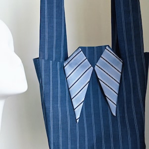 May include: A dark blue tote bag featuring a white pinstripe pattern and two long shoulder straps. The front of the bag is decorated with a pointed collar detail made from light blue and white striped fabric, resembling a dress shirt collar.