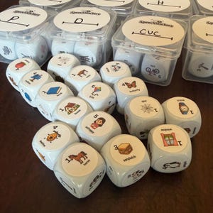 May include: A collection of white plastic dice featuring colorful illustrations of objects like a horse, sandwich, and butterfly, each labeled with a number and word. Several dice are stored in clear plastic containers labeled SpeechStackers.