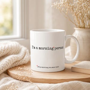 May include: A white ceramic mug featuring black typewriter-style text that reads, "I'm a morning person" with a red asterisk, and smaller text below that reads, "*If by morning you mean 11:30" with a red asterisk.
