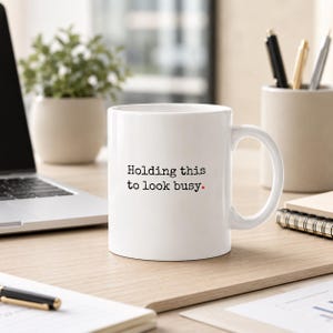 Op de afbeelding: Een witte keramische mok met een handvat, voorzien van de zwarte tekst "Holding this to look busy." met een kleine rode stip aan het einde van de zin. De mok heeft een glad, cilindervormig lichaam en een C-vormig handvat aan de rechterkant.
