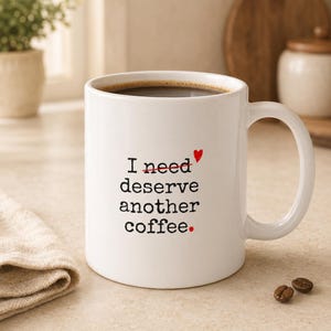 Op de afbeelding: Een witte keramische koffiemok met zwarte tekst in typemachine-stijl die luidt I need deserve another coffee. Het woord need is doorgestreept met een rode lijn, met een klein rood hartje boven de tekst en een rode punt aan het einde.