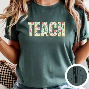 Pu&ograve; includere: Una t-shirt a maniche corte verde scuro con la parola TEACH stampata in un carattere in grassetto. Le lettere sono riempite con un colorato motivo floreale composto da piccoli fiori rosa, gialli e bianchi con foglie verdi su sfondo crema.