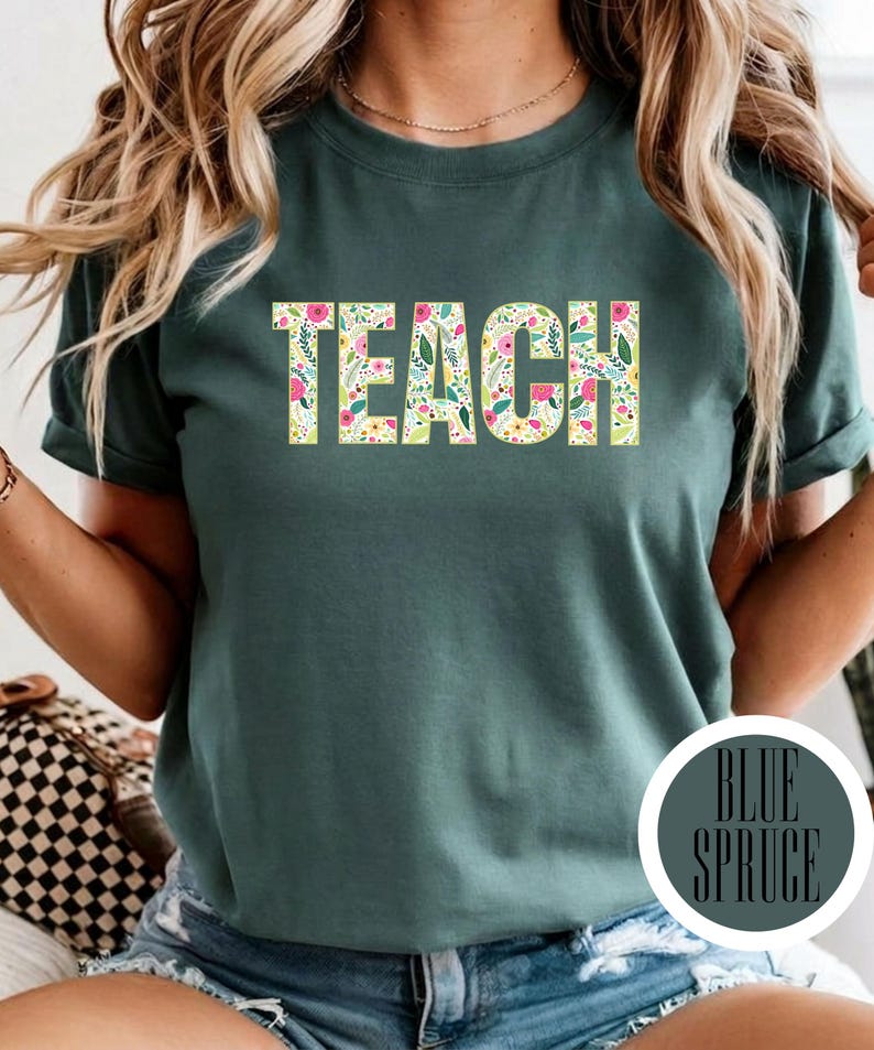 Pu&ograve; includere: Una t-shirt a maniche corte verde scuro con la parola TEACH stampata in un carattere in grassetto. Le lettere sono riempite con un colorato motivo floreale composto da piccoli fiori rosa, gialli e bianchi con foglie verdi su sfondo crema.