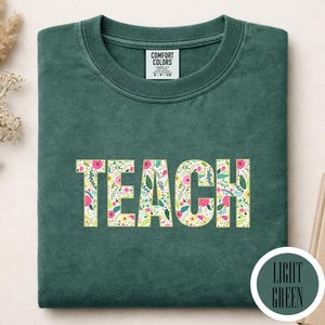 Pu&ograve; includere: Una t-shirt a girocollo verde scuro con la parola TEACH stampata in grandi lettere maiuscole. Le lettere presentano un motivo floreale colorato con elementi botanici rosa, gialli e verdi. Un'etichetta Comfort Colors &egrave; visibile sul colletto.