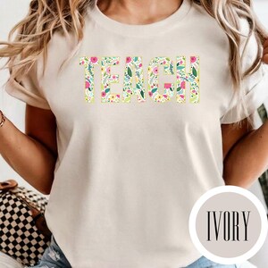 Pu&ograve; includere: Una t-shirt a maniche corte color avorio con la parola TEACH stampata in grandi lettere maiuscole. Le lettere sono riempite con un colorato motivo floreale composto da fiori rosa, gialli e blu mescolati a foglie verdi su sfondo bianco.