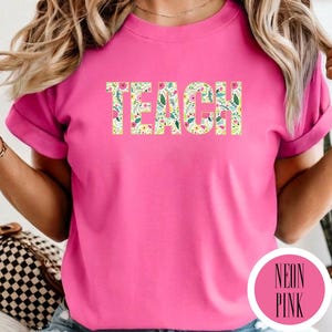 Pu&ograve; includere: Una t-shirt rosa neon con la parola TEACH stampata in grandi lettere maiuscole sul petto. Le lettere sono riempite con un colorato motivo floreale composto da piccoli fiori rosa, gialli e bianchi con foglie verdi su uno sfondo bianco.