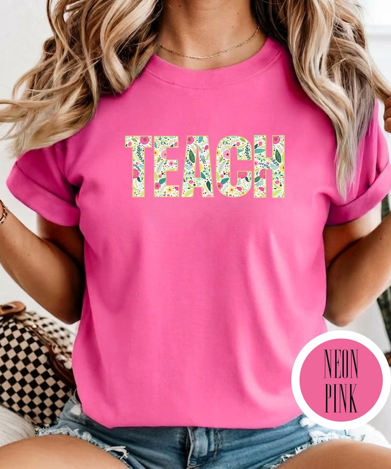 Pu&ograve; includere: Una t-shirt rosa neon con la parola TEACH stampata in grandi lettere maiuscole sul petto. Le lettere sono riempite con un colorato motivo floreale composto da piccoli fiori rosa, gialli e bianchi con foglie verdi su uno sfondo bianco.