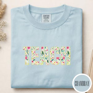 Pu&ograve; includere: Una t-shirt girocollo azzurro chiaro con la parola TEACH stampata in grandi lettere maiuscole sul petto. Le lettere presentano un motivo floreale colorato con elementi botanici rosa, gialli e verdi. Un'etichetta Comfort Colors si trova sul colletto.