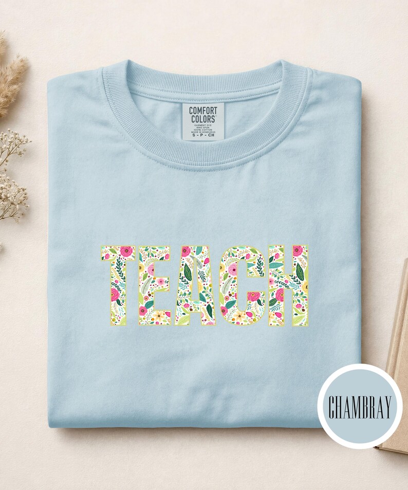Pu&ograve; includere: Una t-shirt girocollo azzurro chiaro con la parola TEACH stampata in grandi lettere maiuscole sul petto. Le lettere presentano un motivo floreale colorato con elementi botanici rosa, gialli e verdi. Un'etichetta Comfort Colors si trova sul colletto.