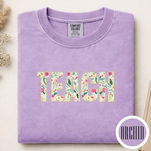 Pu&ograve; includere: Una t-shirt girocollo color lavanda con la parola TEACH stampata in un motivo floreale con elementi botanici rosa, gialli e verdi. La maglietta presenta un'etichetta Comfort Colors sul colletto e un badge circolare con la scritta ORCHID.