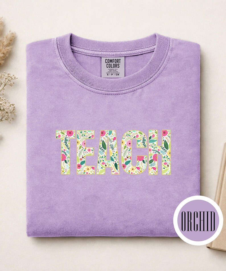 Pu&ograve; includere: Una t-shirt girocollo color lavanda con la parola TEACH stampata in un motivo floreale con elementi botanici rosa, gialli e verdi. La maglietta presenta un'etichetta Comfort Colors sul colletto e un badge circolare con la scritta ORCHID.