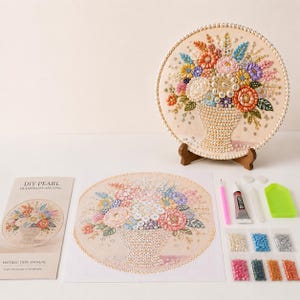 May include: A DIY pearl diamond painting kit featuring a circular canvas with a floral bouquet design, a finished circular artwork on a wooden stand, an instruction manual, a pink applicator tool, glue, and various colored beads.