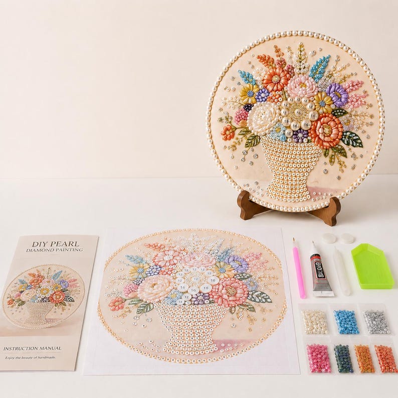 May include: A DIY pearl diamond painting kit featuring a circular canvas with a floral bouquet design, a finished circular artwork on a wooden stand, an instruction manual, a pink applicator tool, glue, and various colored beads.