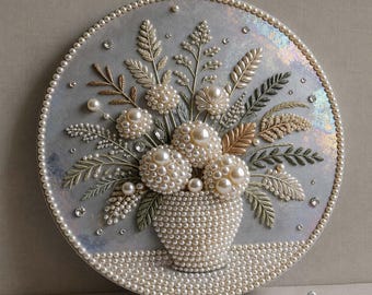 May include: A circular wall art piece featuring a bouquet of white pearl-encrusted flowers in a matching pearl vase. The background is a shimmering, iridescent gray surface. The entire composition is bordered by a single row of small white pearls.