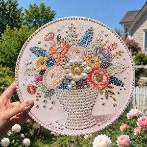 May include: A circular embroidery hoop art piece featuring a bouquet of flowers in a vase. The design is embellished with white pearls, colorful beads, and sequins in shades of pink, yellow, blue, and purple on a light fabric background.