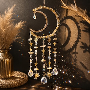 May include: A hanging crescent moon ornament featuring gold-toned metal, embedded with yellow crystal chips, and adorned with dangling chains. The chains include small gold hummingbird and star charms, clear crystal beads, and faceted glass prisms.