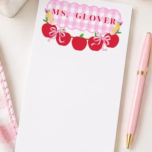 Peut inclure: Un bloc-notes rectangulaire blanc avec une bordure festonnée à carreaux roses en haut et le texte MS. GLOVER en rouge. Le motif comprend trois pommes rouges, deux crayons jaunes et deux nœuds roses disposés en une rangée horizontale décorative.