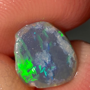 May include: A translucent, irregularly shaped opal gemstone displaying vibrant flashes of neon green and blue light against a milky, semi-transparent base. The stone exhibits a natural, unpolished texture with internal colour play and light reflections.